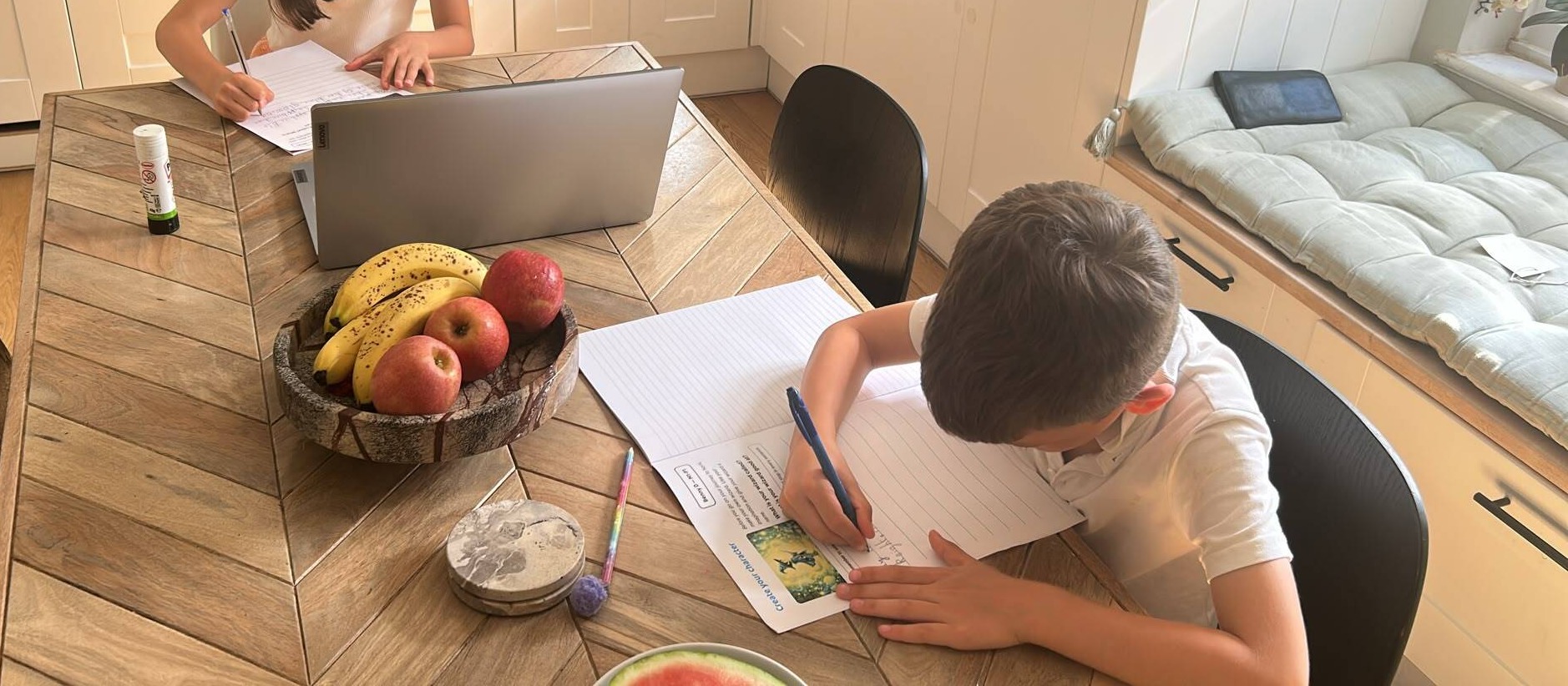 Two children writing by hand on lined paper at a kitchen table, with a laptop open beside them